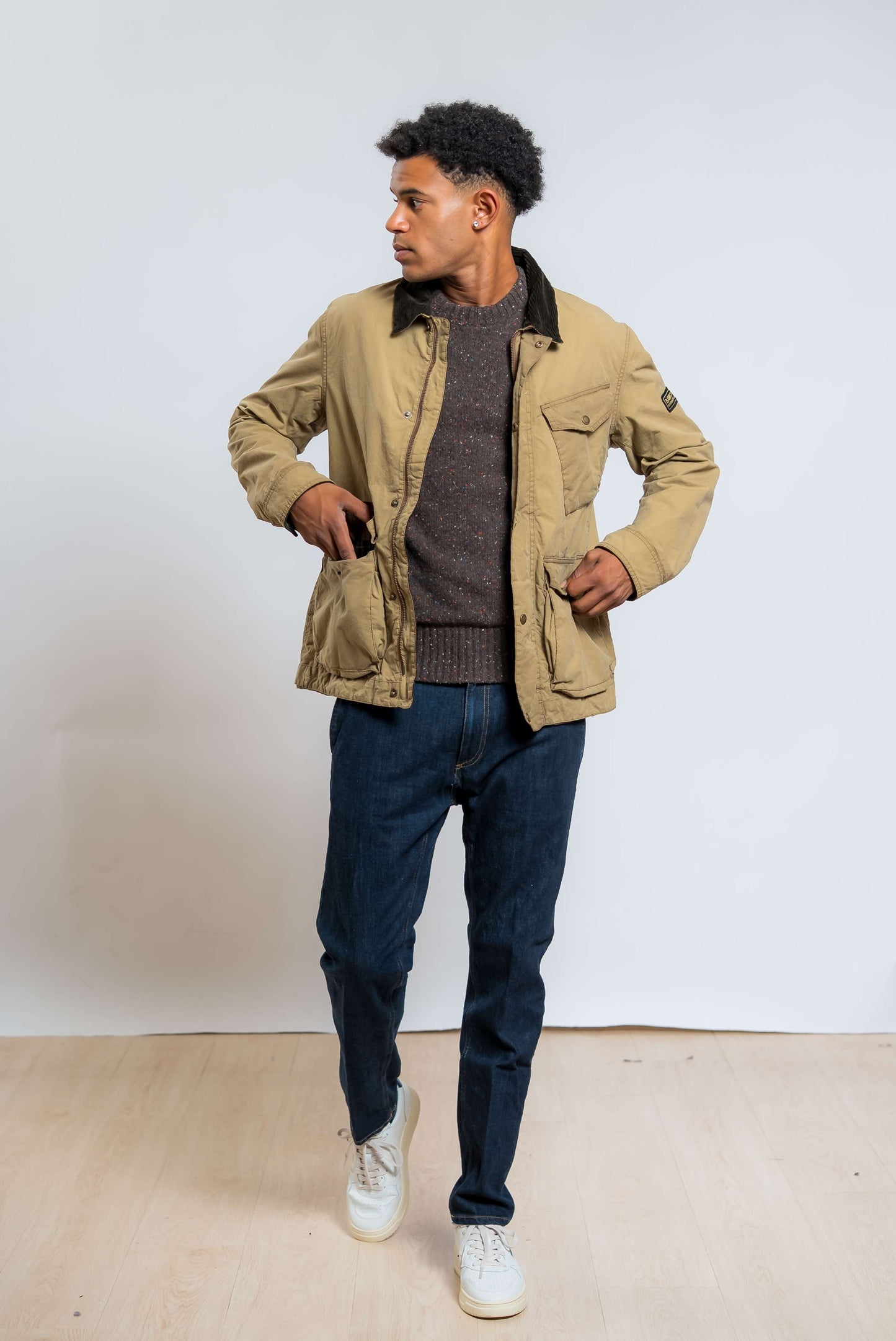 giubbino drifter beige in cotone cerato di barbour international abbinato ad un maglione in lana boucle marrone e un jeans scuro