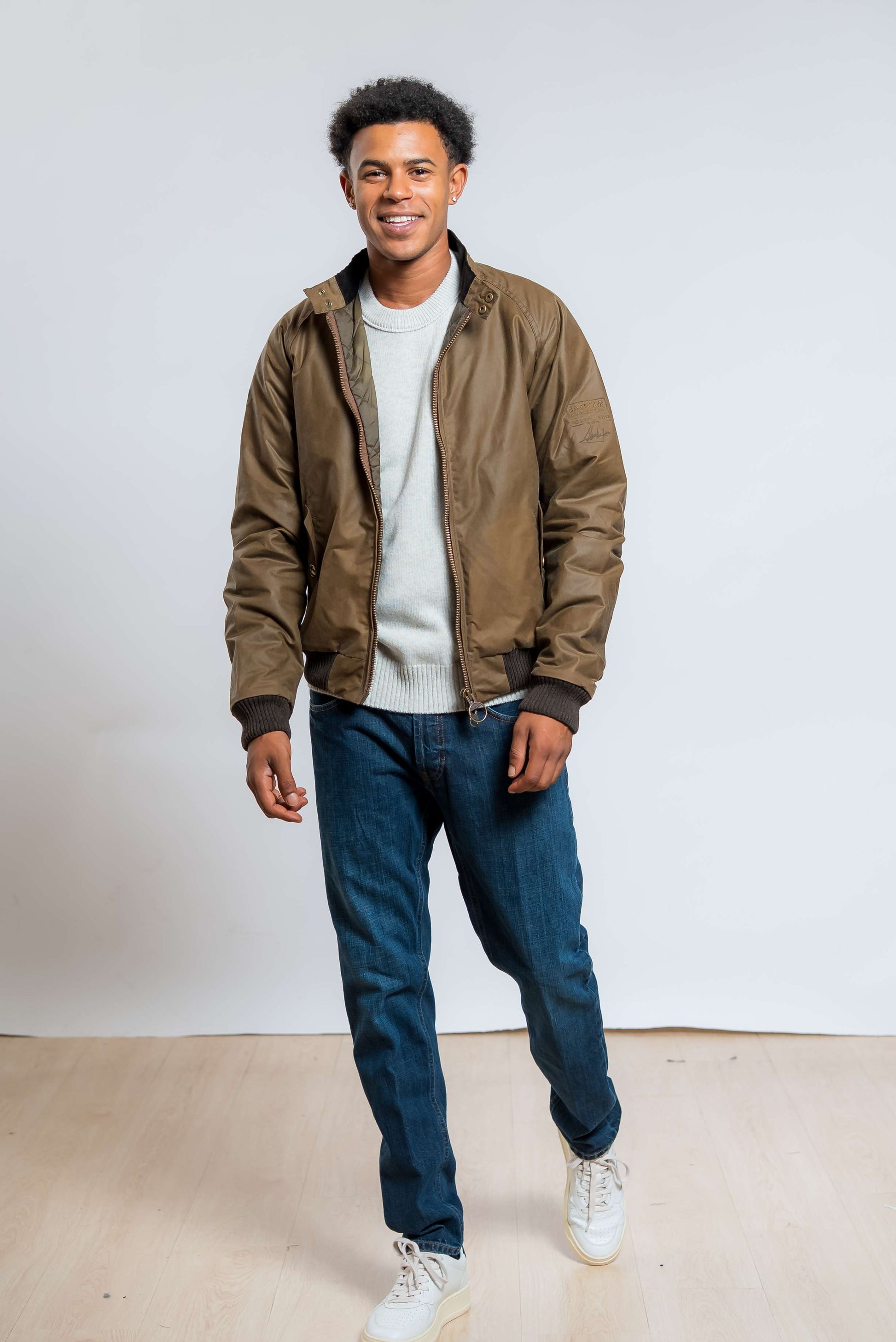 barbour international giubbino modello bomber merchant waxed marrone in cotone cerato abbinato ad un maglione grigio e jeans blu